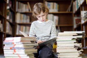 boy reading stacks of books in library - Ранее развитие детей: утомленные детством